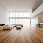 A wide-angle architectural photo of a luxurious open-concept living room featuring continuous wide-plank light oak flooring that seamlessly connects the living area to the adjacent space, illustrating structural design intended to expand the room’s perceived footprint.