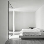 A small, minimalist bedroom painted entirely in a seamless soft white, where the ceiling and walls blend together, featuring a large floor-to-ceiling mirror to create the illusion of expanded space and increased height.