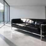 A modern minimalist living room featuring a black leather Le Corbusier style sofa with a visible chrome steel frame as the architectural centerpiece.