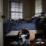 A realistic photograph of a severely cluttered, unmade private bedroom with large piles of laundry, disorganized surfaces, and poor lighting, conveying visual stress and anxiety.