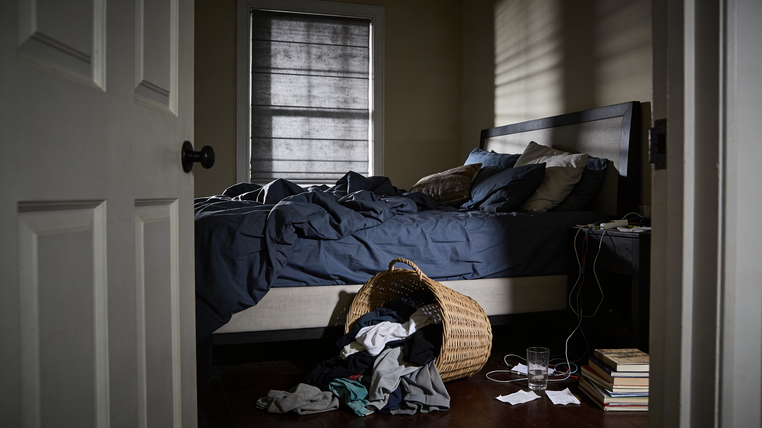A realistic photograph of a severely cluttered, unmade private bedroom with large piles of laundry, disorganized surfaces, and poor lighting, conveying visual stress and anxiety.